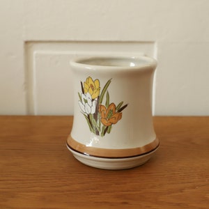 Pode incluir: Um porta-escovas de dentes de cerâmica branca com uma borda marrom e um design floral com crocus amarelos, brancos e laranja.