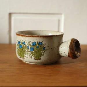 Könnte beinhalten: Eine weiße Suppentasse aus Keramik mit braunem Rand und einem Blumendesign mit blauen Blüten und grünen Blättern. Die Tasse hat einen Henkel, der seitlich herausragt.