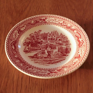 Op de afbeelding: Een wit keramiek bord met een rood en wit bloemenpatroon. Het bord toont een scène met een huis, een tuin en een hek.