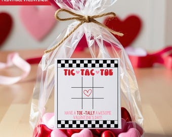 Op de afbeelding: Een printbare Valentijnskaart met een tic-tac-toe spel en de tekst "Have a toe-tally awesome Valentine's Day!" De kaart zit in een doorzichtige plastic zak met een bruine touw strik en roze hartvormige snoepjes.