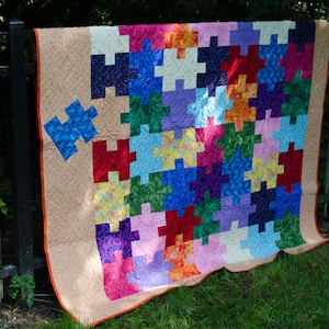 May include: A colorful quilt made of fabric puzzle pieces in shades of red, blue, green, purple, orange, and yellow. The quilt is hanging on a black metal fence.