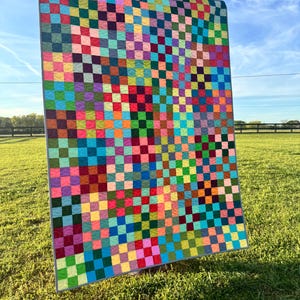 May include: A large, colorful quilt with a checkerboard pattern of various colors, including pink, green, blue, and yellow. The quilt is displayed outdoors on a sunny day, with a green grassy background and a blue sky.