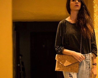 Pode incluir: Uma mulher vestindo uma camisa de manga comprida cinza escura e jeans azul claro com detalhes desgastados. Ela está carregando uma bolsa de embreagem de cortiça com uma alça de corrente dourada.