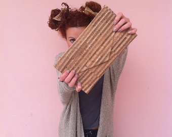 May include: A woman with red hair is holding a cork clutch in front of her face. The clutch is brown and has a striped pattern. She is wearing a light gray cardigan and dark jeans.