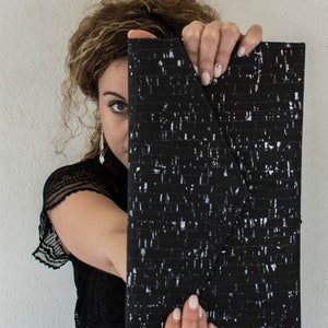 Pode incluir: Uma carteira de m&atilde;o preta e branca com um padr&atilde;o e um fecho de aba. A carteira de m&atilde;o &eacute; segurada na frente do rosto de uma pessoa.
