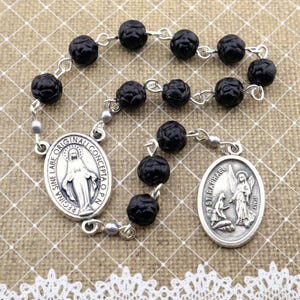 Peut inclure: Un chapelet avec des perles noires texturées et des maillons en métal argenté. Le chapelet comporte deux pendentifs ovales avec des images et du texte religieux. Le fond est un tissu beige avec une bordure en dentelle blanche.