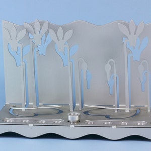 May include: Silver menorah with a floral design. The menorah has nine candle holders and a central shamash holder. The floral design is cut out of the metal and creates a delicate and elegant look.