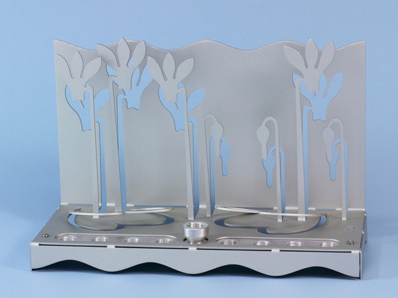 May include: Silver menorah with a floral design. The menorah has nine candle holders and a central shamash holder. The floral design is cut out of the metal and creates a delicate and elegant look.