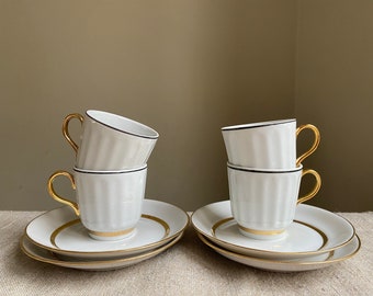May include: Set of four white porcelain teacups with gold trim and black accents. The cups are stacked in pairs on matching saucers.