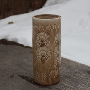 K&ouml;nnte beinhalten: Eine braune Keramikvase mit einem geometrischen Muster aus Kreisen und Linien. Die Vase steht auf einer Holzoberfl&auml;che.