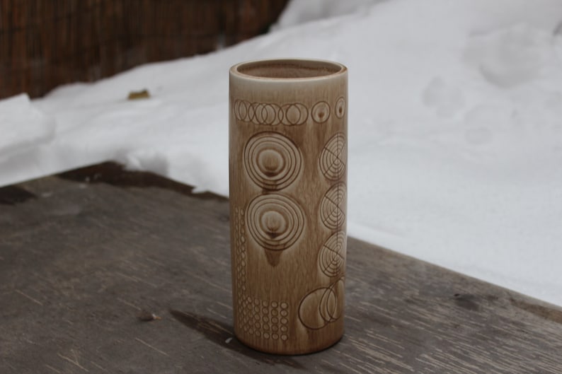 K&ouml;nnte beinhalten: Eine braune Keramikvase mit einem geometrischen Muster aus Kreisen und Linien. Die Vase steht auf einer Holzoberfl&auml;che.