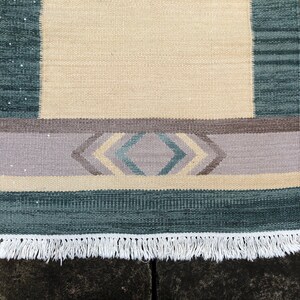 May include: A woven rug with a cream background, a light purple band with a geometric pattern in shades of blue and cream, and a dark blue border with a white fringe.