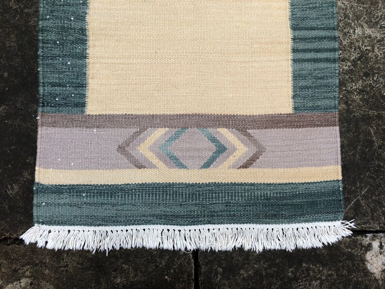 May include: A woven rug with a cream background, a light purple band with a geometric pattern in shades of blue and cream, and a dark blue border with a white fringe.