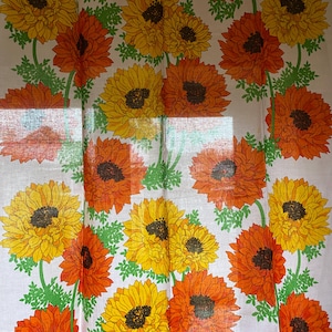 May include: White fabric with a repeating pattern of orange and yellow flowers. The flowers have a dark brown center and are surrounded by green leaves.