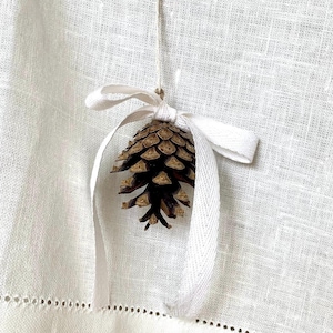 Puede incluir: Un cono de pino marrón atado con una cinta blanca, colgando de un fondo de lino blanco.