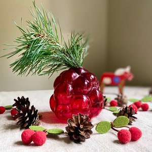 Puede incluir: Un jarrón de vidrio rojo con forma de racimo de bayas con una rama de agujas de pino verdes en su interior. El jarrón está rodeado de pequeñas piñas y bolas de fieltro rojas.