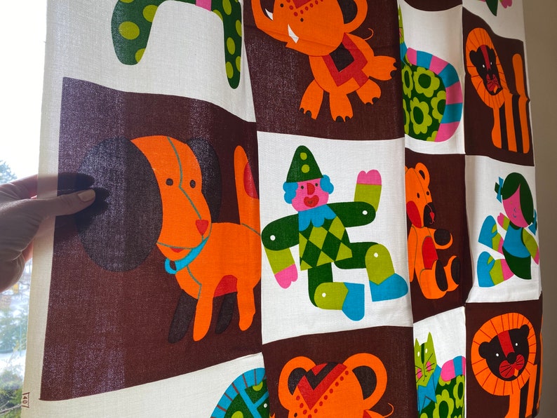 May include: A brown and white fabric with a colorful pattern of animals and a clown. The animals include a dog, a lion, an elephant, a cat, and a bear. The clown is wearing a green and yellow suit with a red hat.