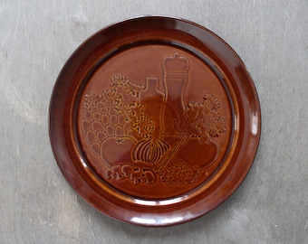 May include: A brown ceramic plate with a raised design of vegetables, herbs, and a bottle and pepper shaker. The plate has a brown glaze.