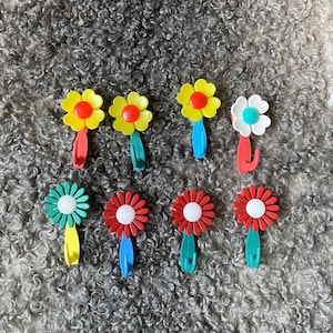 Könnte beinhalten: Acht bunte, blumenförmige Haken in einem gelben, roten und blauen Farbschema. Die Haken sind aus Kunststoff und haben ein verspieltes Design.
