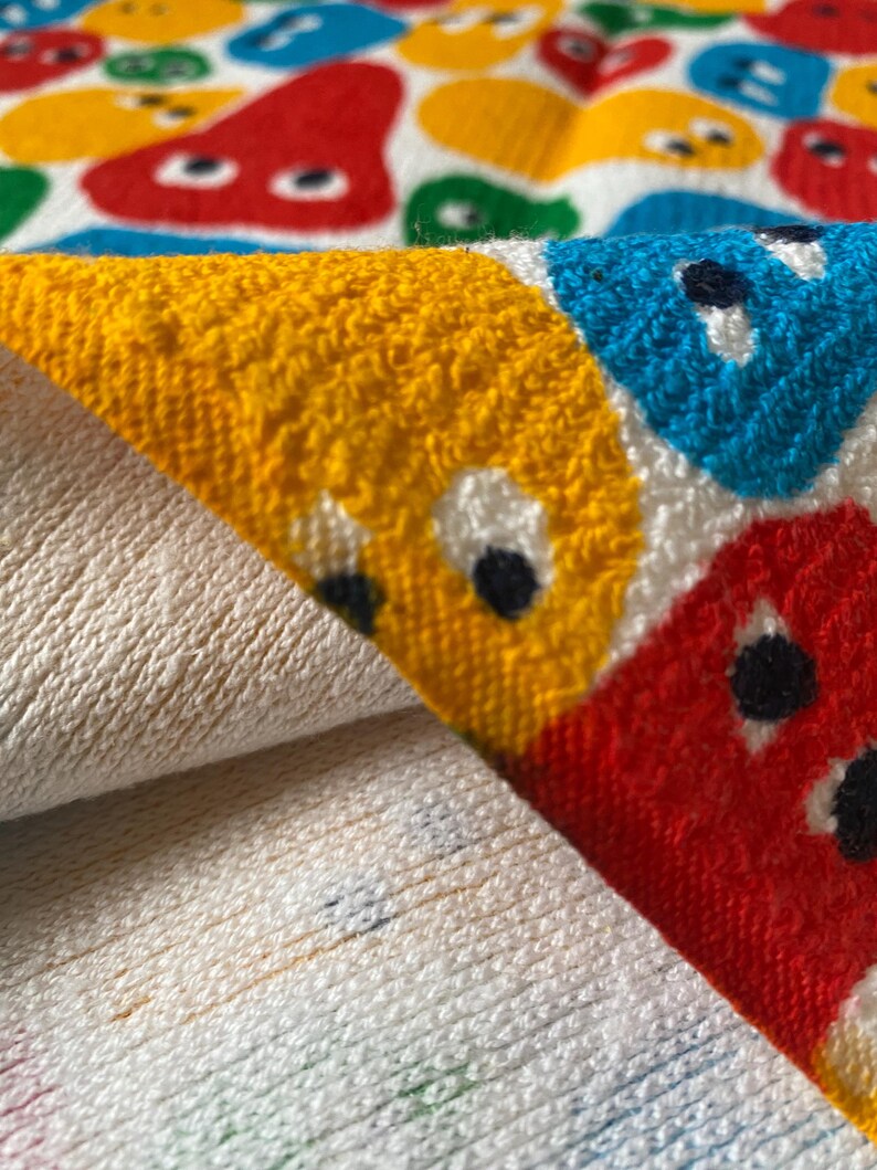 Pu&ograve; includere: Un asciugamano bianco piegato con un motivo colorato di facce di cartoni animati in rosso, giallo, blu e verde. La trama dell'asciugamano &egrave; visibile, con un aspetto morbido e assorbente. Il design &egrave; giocoso e adatto a un bagno o alla spiaggia.