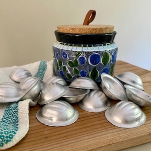 Peut inclure: Un pot en céramique avec un couvercle en liège et une tirette en cuir, décoré de motifs floraux bleus et verts. Plusieurs petits moules en métal argenté sont éparpillés, probablement pour la pâtisserie. Les objets reposent sur une surface en bois.