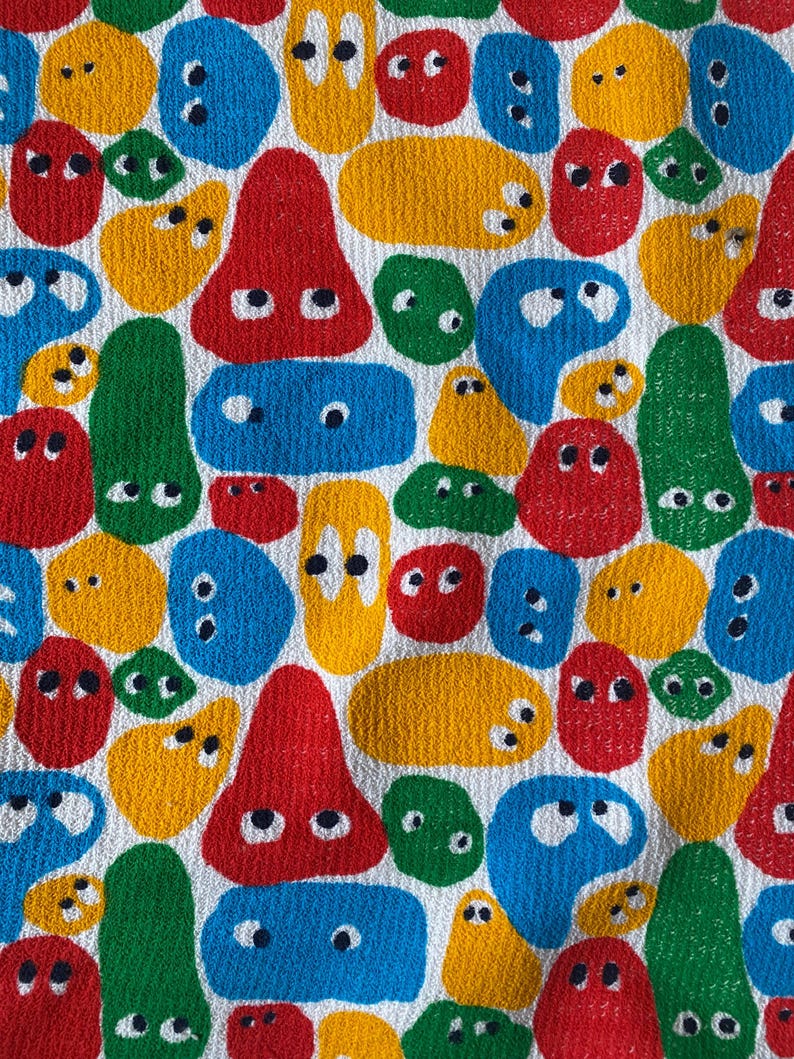 Pu&ograve; includere: Motivo tessile colorato con varie forme astratte rosse, gialle, blu e verdi, ciascuna con due piccoli dettagli neri a forma di occhi. Le forme sono disposte su uno sfondo bianco, creando un design giocoso e vivace.