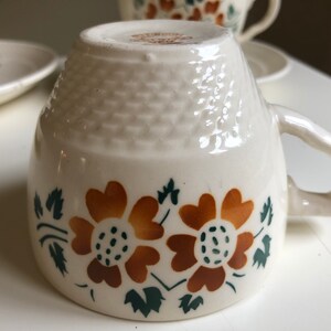 Peut inclure: Une tasse &agrave; th&eacute; en c&eacute;ramique blanche avec un motif floral brun et vert. La tasse a un fond textur&eacute; et une anse.