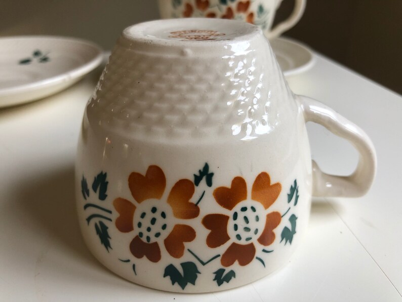 Peut inclure: Une tasse &agrave; th&eacute; en c&eacute;ramique blanche avec un motif floral brun et vert. La tasse a un fond textur&eacute; et une anse.