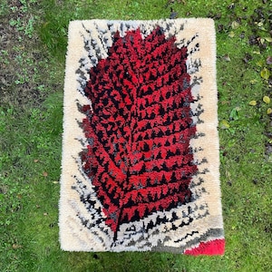 May include: Rectangular rug with a central red and black leaf design. The leaf is surrounded by a cream-colored border with black and gray accents. The rug is placed on green grass.