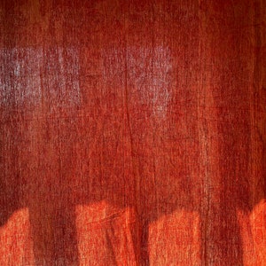 May include: A red fabric curtain with a textured woven pattern. The curtain is hanging in a window and is illuminated by sunlight, creating shadows on the fabric.