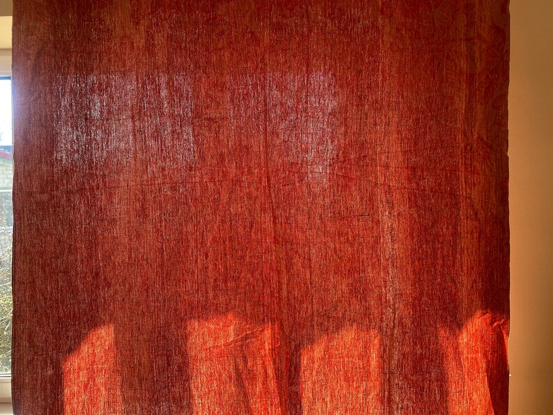 May include: A red fabric curtain with a textured woven pattern. The curtain is hanging in a window and is illuminated by sunlight, creating shadows on the fabric.