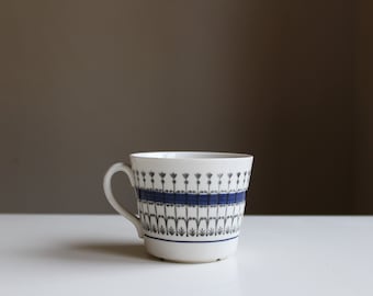 Puede incluir: Una taza de té de cerámica blanca con un patrón floral azul y gris. La taza tiene un asa y una forma redondeada.