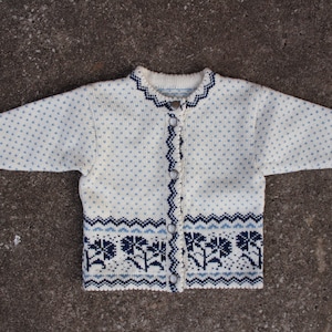 May include: A white knitted cardigan with a blue and white patterned design. The cardigan has a button-up front and long sleeves.