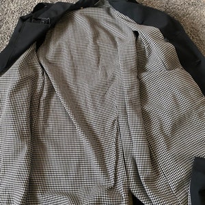 Op de afbeelding: Zwarte trenchcoat met een zwart-witte ruitjesvoering. De jas is open en de voering is zichtbaar.