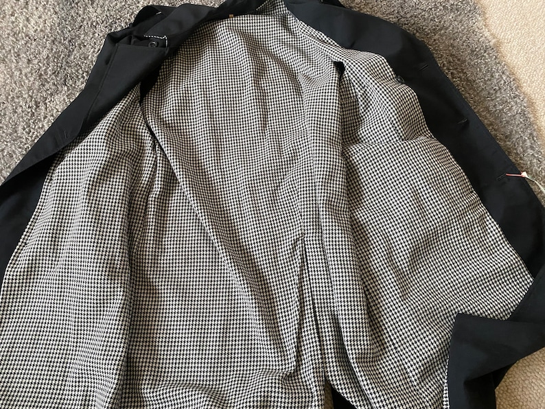 Op de afbeelding: Zwarte trenchcoat met een zwart-witte ruitjesvoering. De jas is open en de voering is zichtbaar.