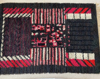 May include: A black and red geometric patterned rug with a shaggy texture. The rug has a central design with a black background and red and white geometric shapes.