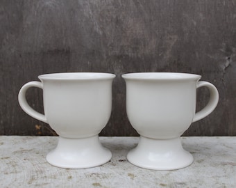 Peut inclure: Deux tasses en céramique blanche avec un piédestal. Les tasses sont vides et ont une finition lisse et brillante.