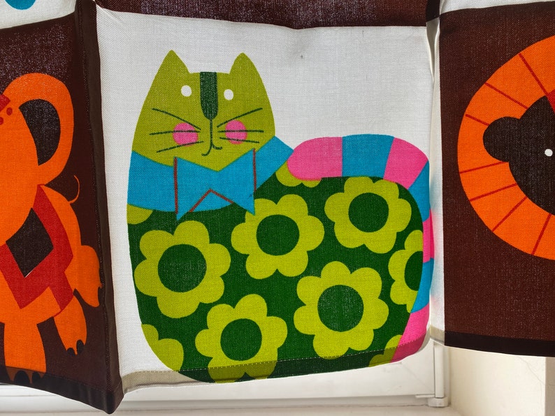 May include: A green cat with a blue bow tie and pink and blue tail, wearing a green and yellow floral pattern. The cat is on a white background.