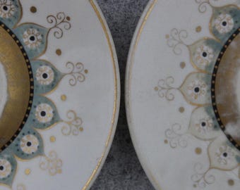 May include: Two off-white plates with gold rims and intricate designs. The plates feature a gold inner circle, teal and gold floral patterns, and small black dots. The design is symmetrical and elegant.