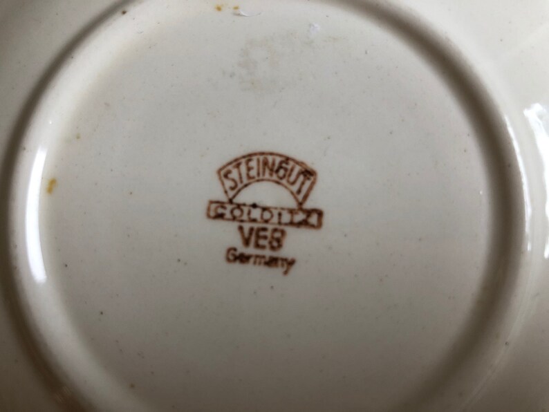 Peut inclure: Gros plan d'une marque en gr&egrave;s brun sur une assiette blanche. La marque indique "Steingut Goldnix VEB Germany".