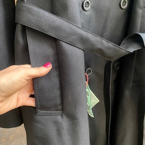 Op de afbeelding: Een zwarte trenchcoat met een dubbele rij knopen en een riem. De jas heeft een zak aan de linkerkant en een label dat aan de riem hangt.