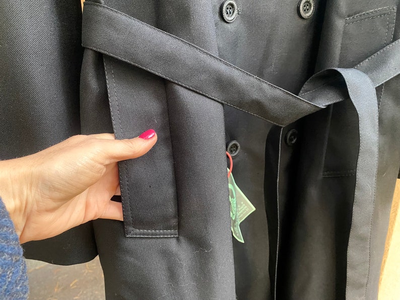 Op de afbeelding: Een zwarte trenchcoat met een dubbele rij knopen en een riem. De jas heeft een zak aan de linkerkant en een label dat aan de riem hangt.