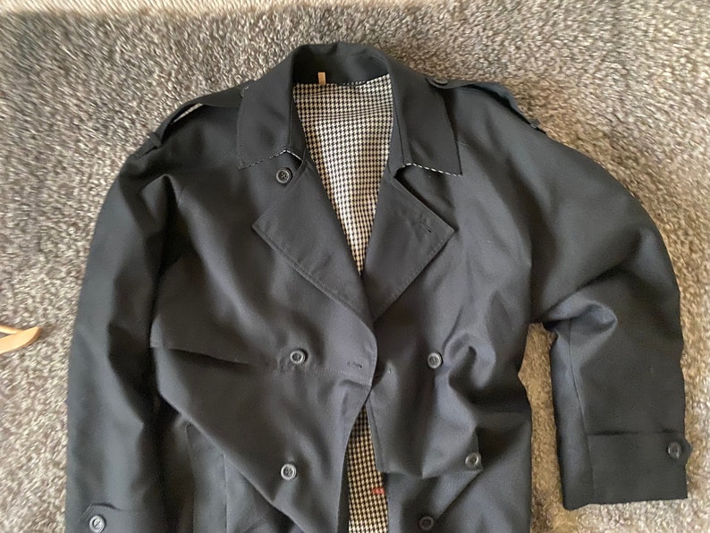Op de afbeelding: Een zwarte dubbelrijdige trenchcoat met een geruite voering.