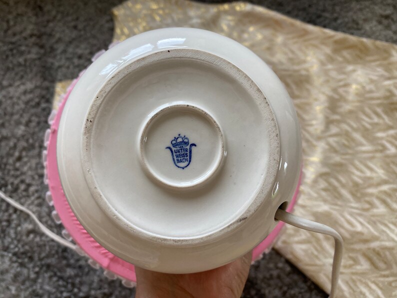 Może przedstawiać: Biała ceramiczna podstawa lampy z niebiesko-białym logo z napisem "Unter Weiss Bach" i r&oacute;żową koronkową ozdobą wok&oacute;ł podstawy.