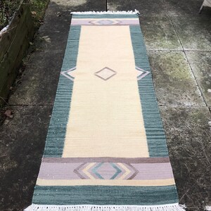 May include: A long, rectangular rug with a cream background and a geometric pattern in shades of green, purple, and pink. The rug has a white fringe on both ends.