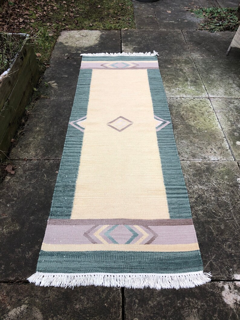 May include: A long, rectangular rug with a cream background and a geometric pattern in shades of green, purple, and pink. The rug has a white fringe on both ends.