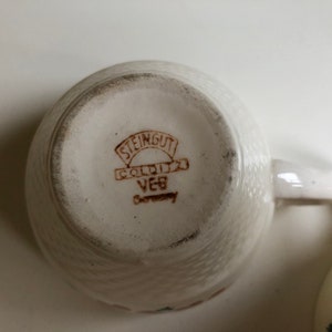 Peut inclure: Gros plan du fond d'une tasse &agrave; th&eacute; en c&eacute;ramique blanche avec un logo brun qui indique "Steingut Colditz VEB Germany".