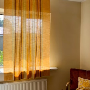 May include: A sheer window curtain featuring vertical stripes in varying shades of yellow, orange, and brown. The fabric has a visible woven texture that allows light to pass through, creating a warm glow across the surface of the material.