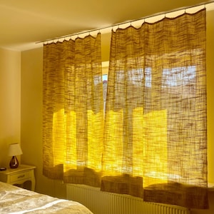 Peut inclure: Deux rideaux transparents brun doré sont suspendus à une tringle argentée, encadrant une fenêtre ensoleillée. Les rideaux ont un aspect texturé et tissé, avec une bordure marron plus foncée en bas. La pièce est éclairée par une lumière naturelle et chaleureuse.