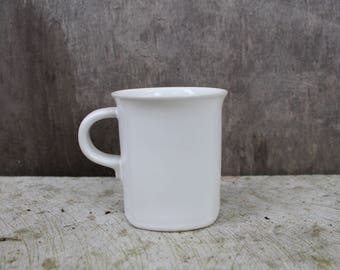 May include: A white ceramic mug with a handle. The mug is empty and sitting on a light brown wooden surface.